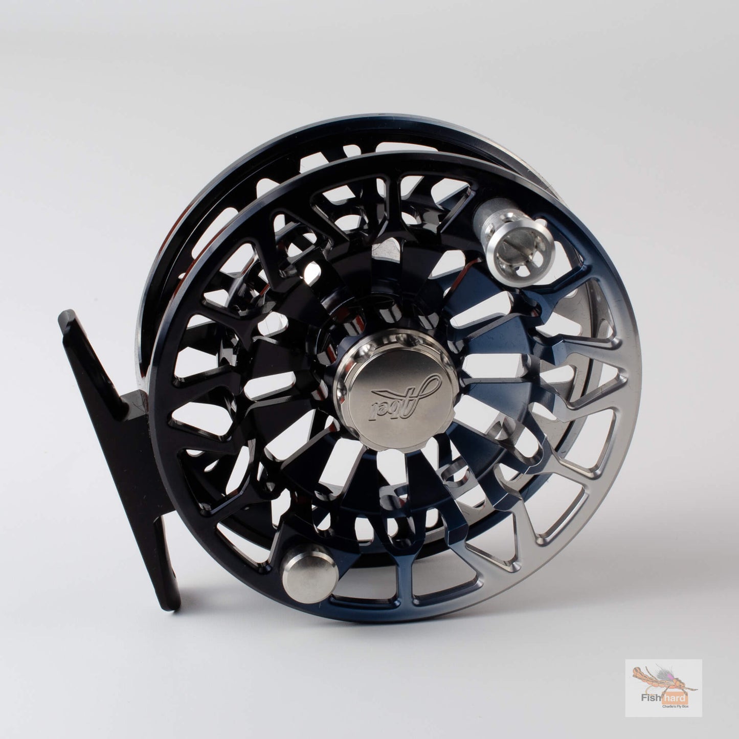 A close-up studio shot of a fly fishing reel with a black body, silver metallic accents, and a black mounting tab, set against a light gray background.