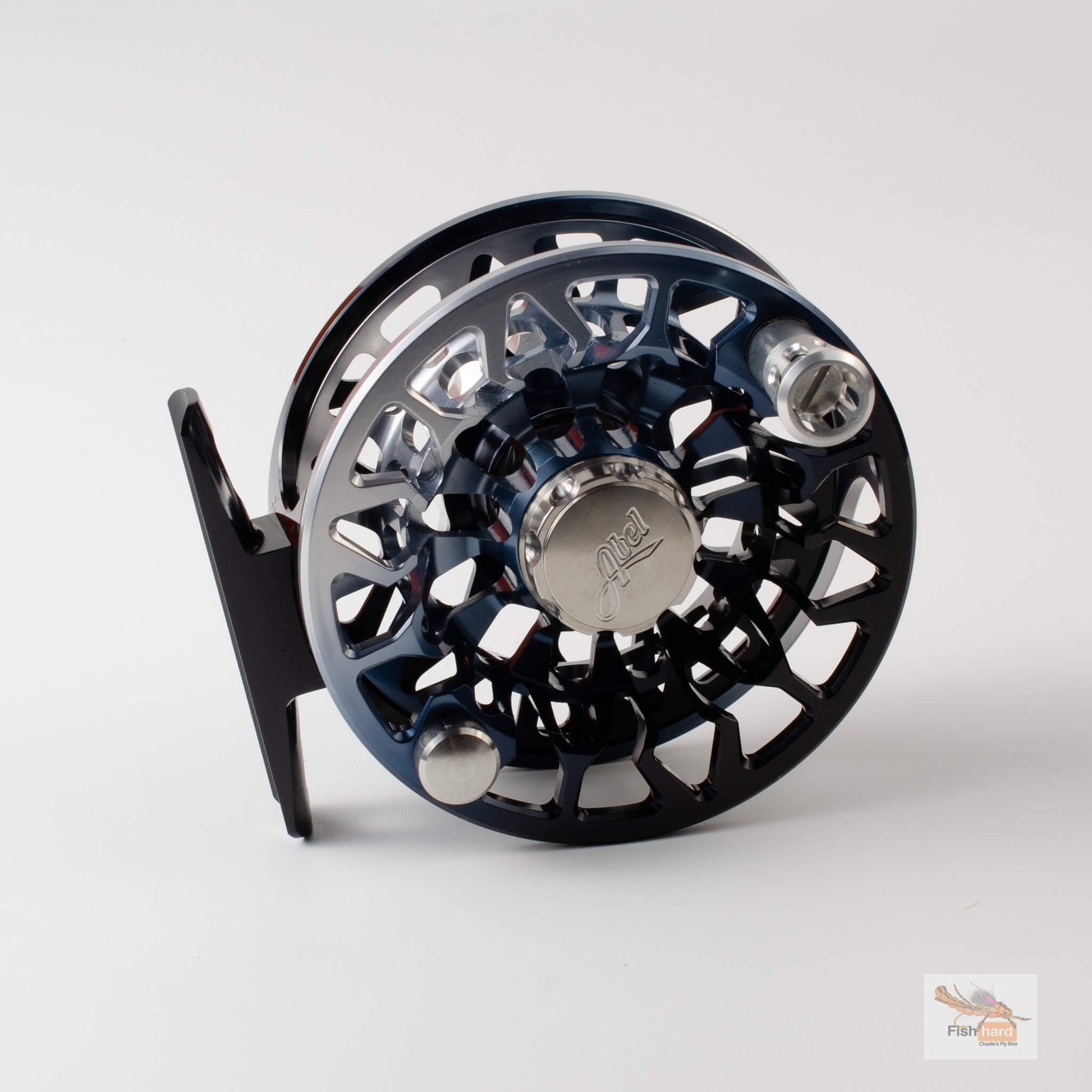 A close-up studio shot of a fly fishing reel, showing its metallic blue and black body, silver spool, and black handle against a light gray background.