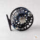 A close-up studio shot of a fly fishing reel, showing its metallic blue and black body, silver spool, and black handle against a light gray background.