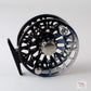 A close-up studio shot of a fly fishing reel with a black body, silver metallic accents, and a black mounting tab, set against a light gray background.