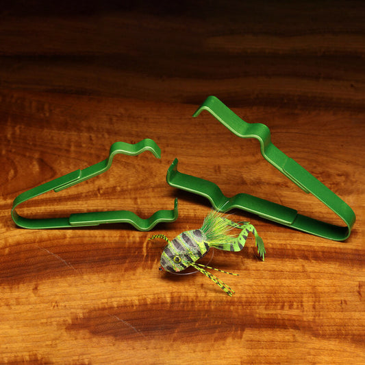 Green metal fly-tying clamps displayed on a wooden surface next to a striped green and yellow nymph lure.